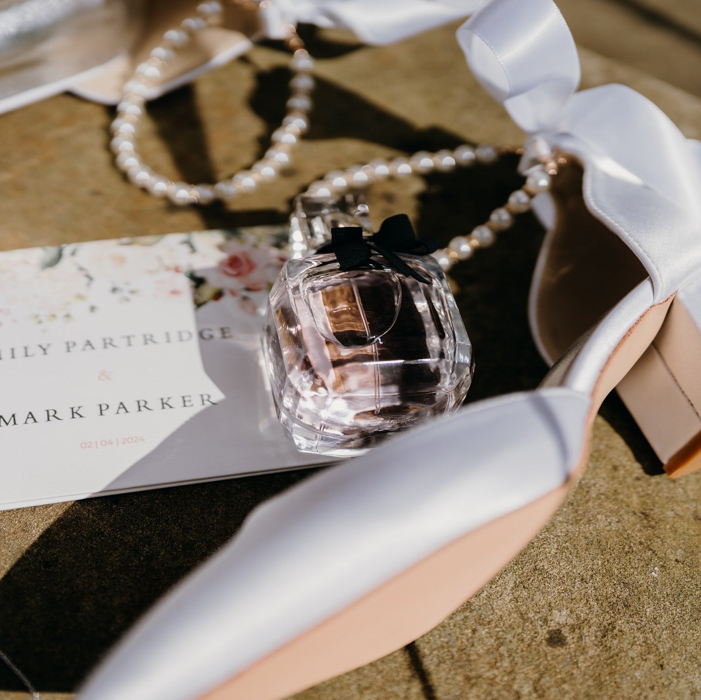 Floral save the date card with wedding details, styled with satin bridal heels, pearl necklace, and pink perfume bottle on a sunlit surface.