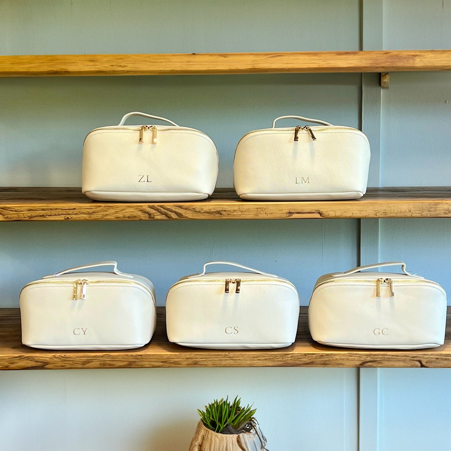 Row of personalised cream vanity cases with gold zips, each monogrammed with initials, displayed on rustic wooden shelves against a pale blue wall.