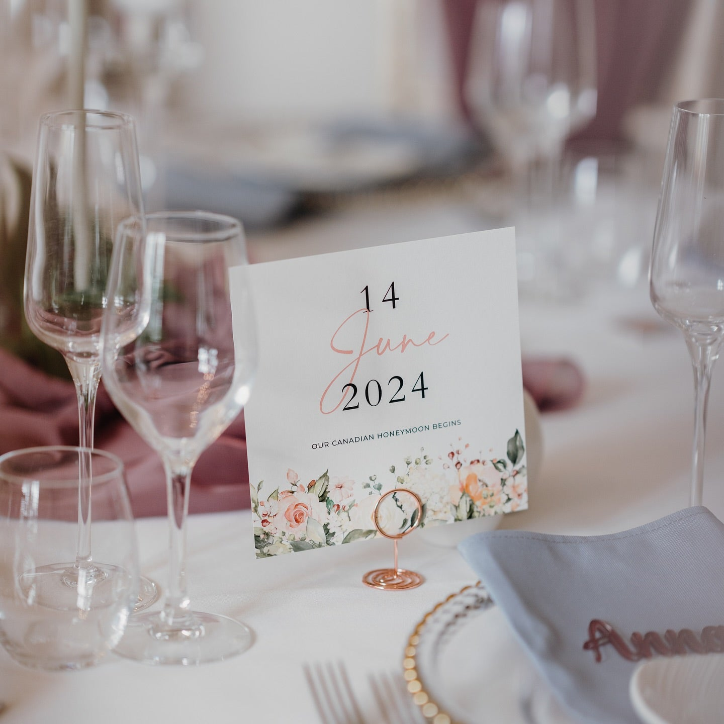 Floral table name card with a custom date design, displayed in a copper holder on a formal wedding table setting with glassware and personalised napkin.