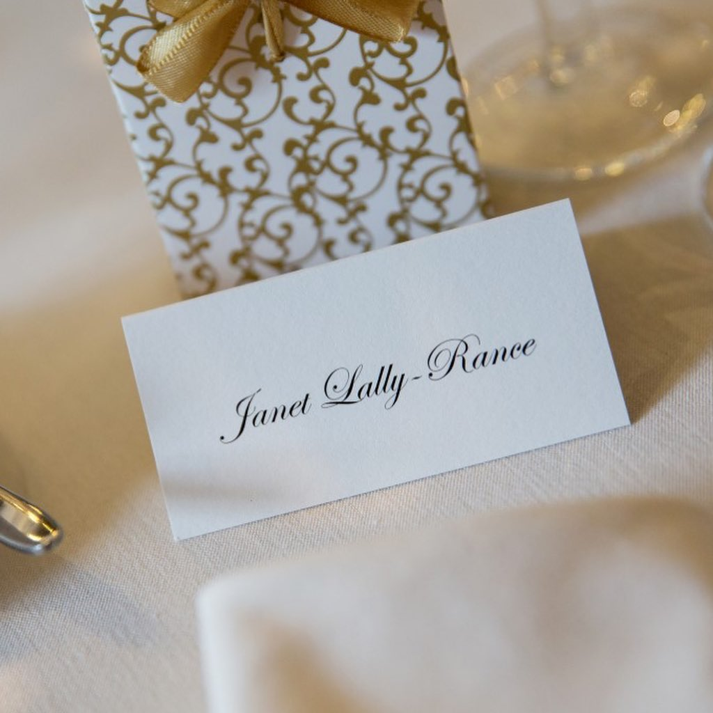 Elegant wedding place card with guest name in script font, displayed on a formal table setting beside a gold-patterned favour box with satin ribbon.
