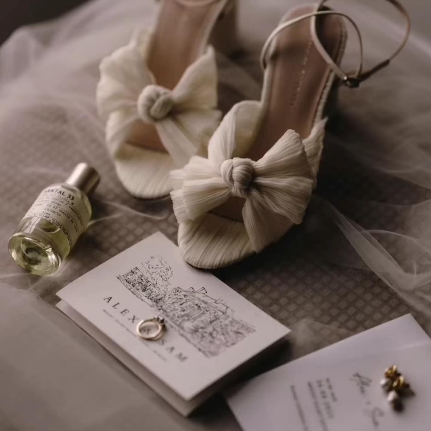 Flat lay featuring knotted ivory bridal heels, hand-drawn wedding invitation with venue sketch, gold ring, perfume bottle, and pearl earrings on tulle fabric.