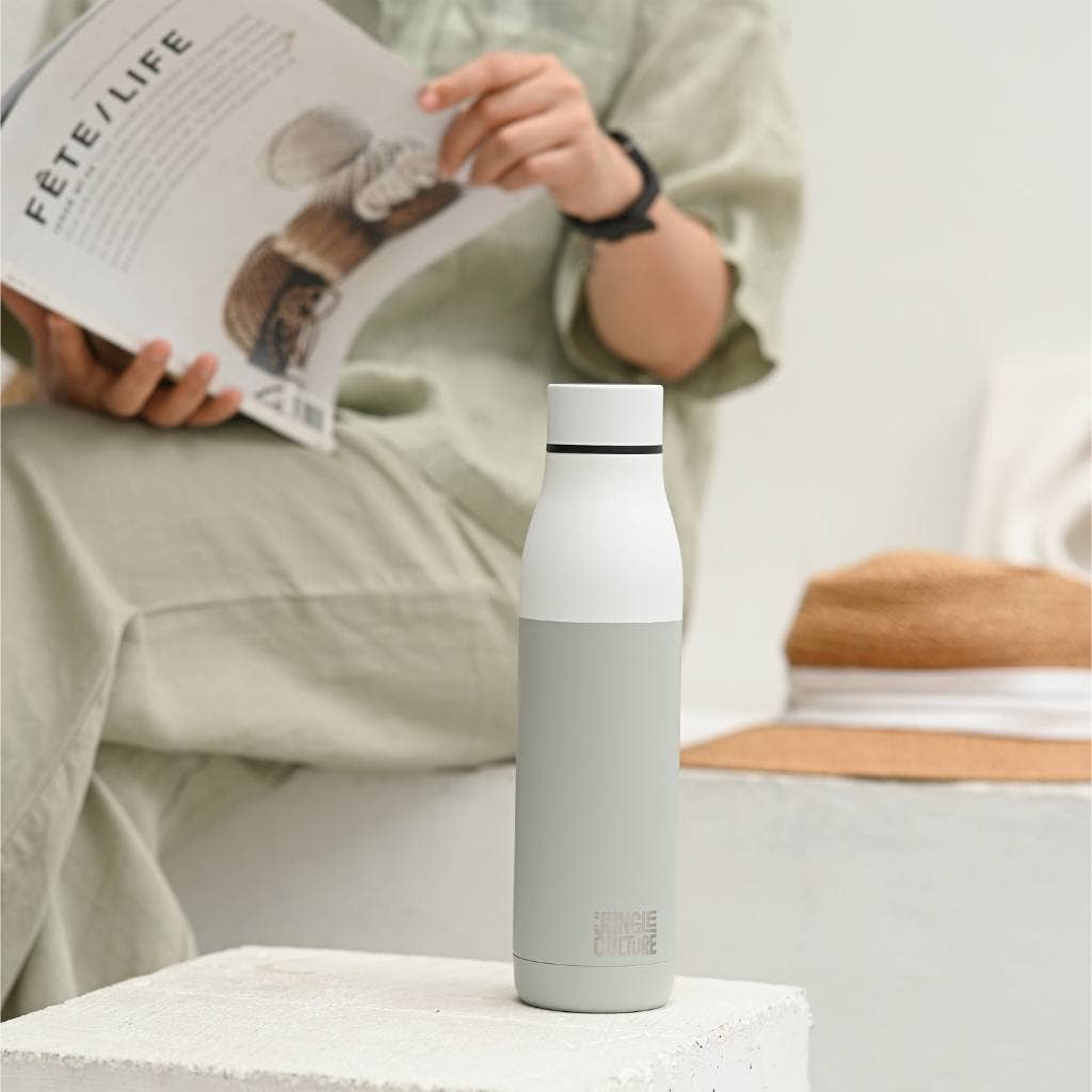 Insulated stainless steel water bottle standing upright on coffee table with person reading magazine behind