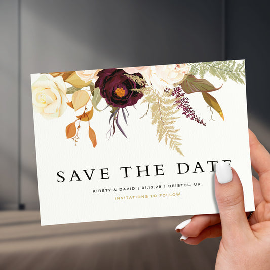 brown and burgundy florals save the date card held in a hand with painted white names, held up against a stylish sleek room background
