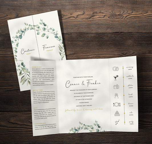 Olive branch gatefold wedding invitation, showing a timeline panel, guest information and the main information. The invitation is set on a table next to props.