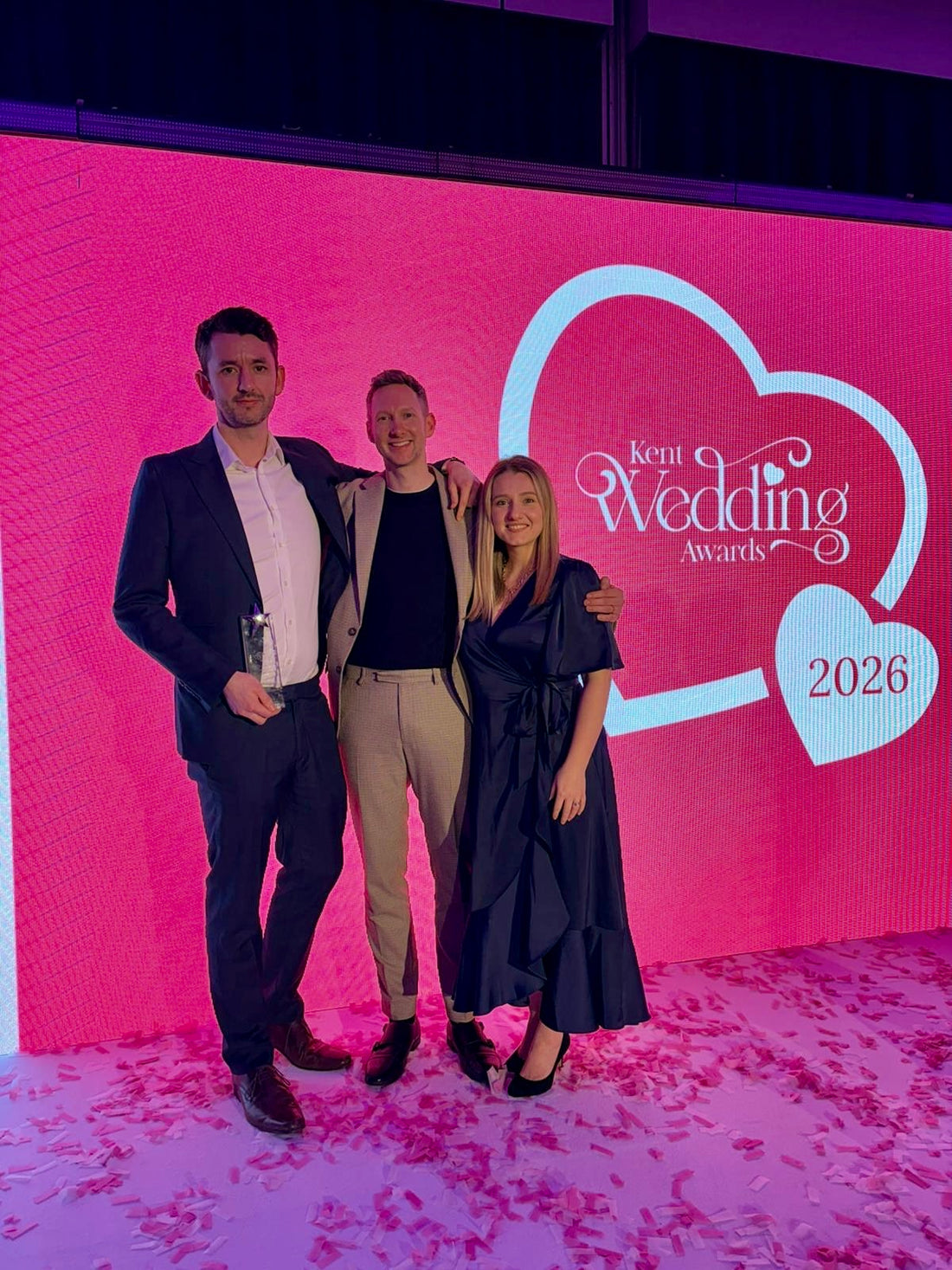 3 people stood holding a wedding award in front of a big pink screen at an event