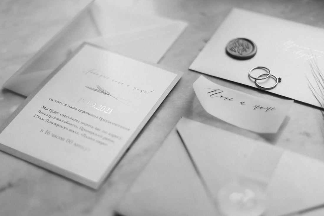 Black and white flat lay of minimalist wedding stationery including invitations, envelopes, a wax seal, and two wedding rings arranged on a marble surface.