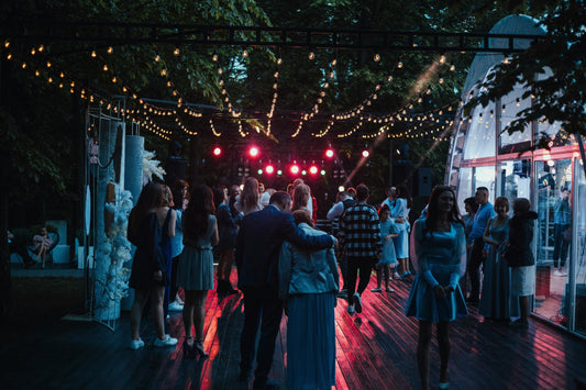 Guests enjoying an outdoor evening wedding reception under string lights, with music, dancing, and a lively atmosphere surrounded by trees.