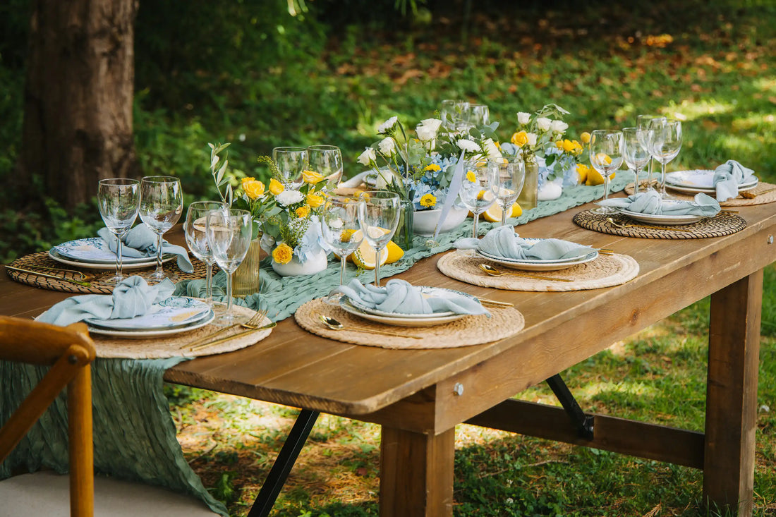 Outdoor wedding reception table set with place settings, folded napkins, glassware and floral centrepieces in a garden setting.