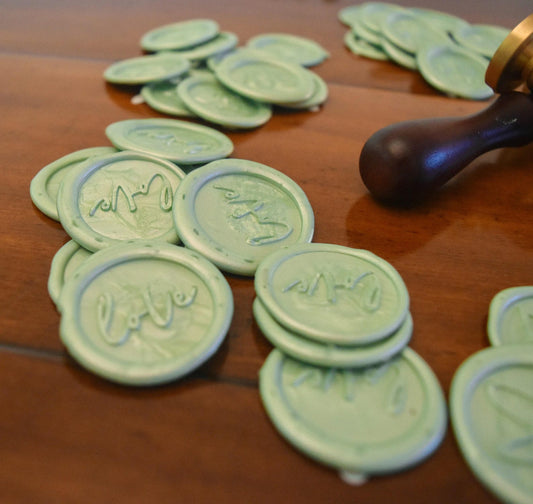 Close-up of sage green self-adhesive wax seals stamped with “love,” displayed on a wooden surface next to a wax seal stamp tool.
