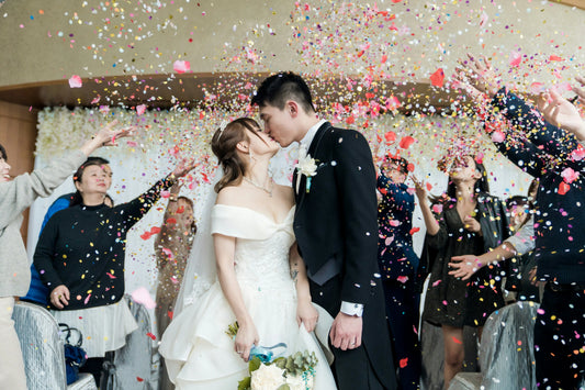 Bride and groom kissing as guests throw colourful confetti during a joyful wedding celebration indoors.