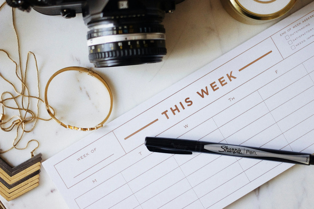 Flat lay of a weekly planner with a camera, gold jewellery, candle and pen arranged on a marble surface, styled as a modern wedding planning workspace.
