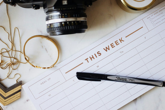 Flat lay of a weekly planner with a camera, gold jewellery, candle and pen arranged on a marble surface, styled as a modern wedding planning workspace.