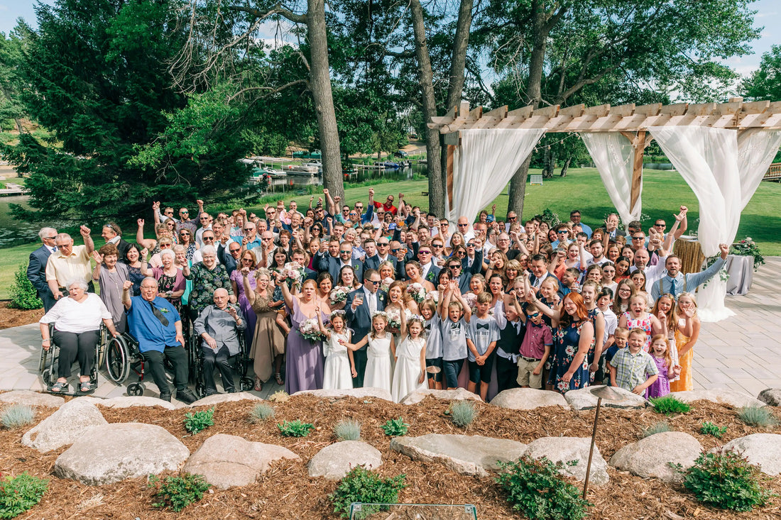Large group of wedding guests celebrating outdoors beneath a wooden pergola, raising their hands in a joyful group photo at a garden wedding venue.