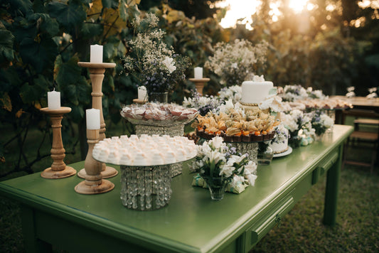 Outdoor sage green wedding reception dessert table with floral arrangements, white candles on wooden holders, cakes, and crystal cake stands at golden hour.