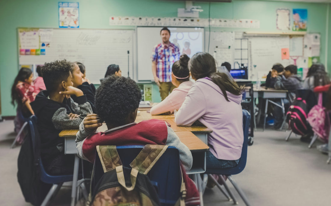 A male teacher in a red checkered shirt standing in front of a class of young teenage pupils. They look on intently and interested and he teaches the class. 