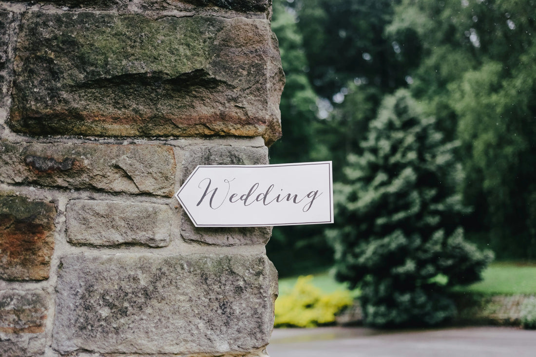 Directional wedding sign reading “Wedding” attached to a stone wall, pointing towards an outdoor venue with greenery in the background.