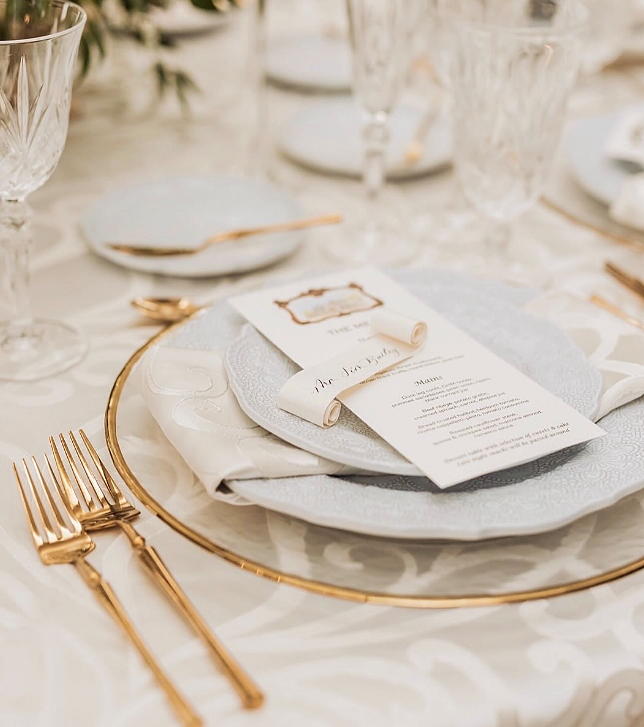 Elegant wedding menu and name scroll displayed on a white and gold place setting with layered plates, gold cutlery, and crystal glassware.