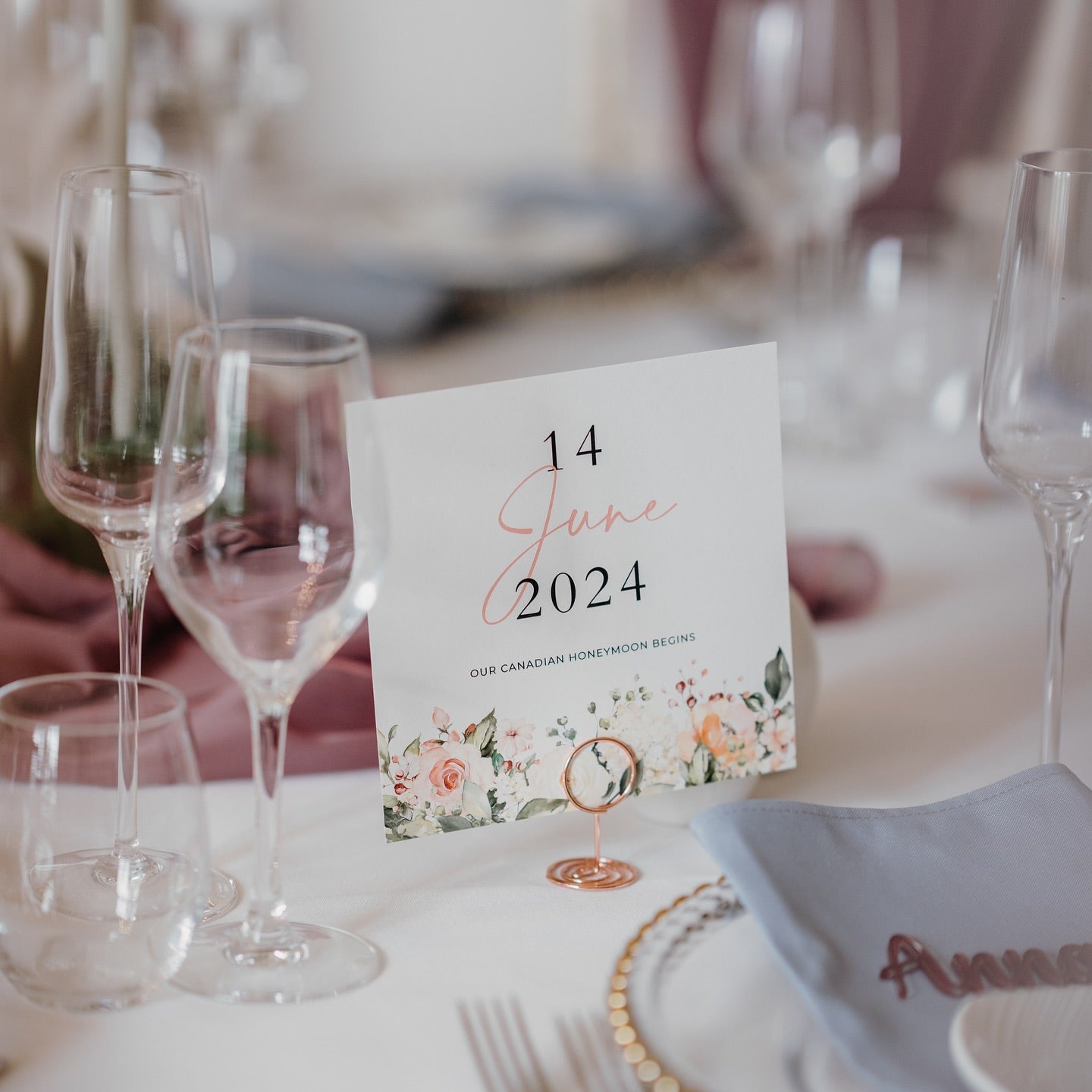 Table card displaying 14 June 2024 with floral border, styled with glassware, blush napkins, and white table setting for a wedding reception.