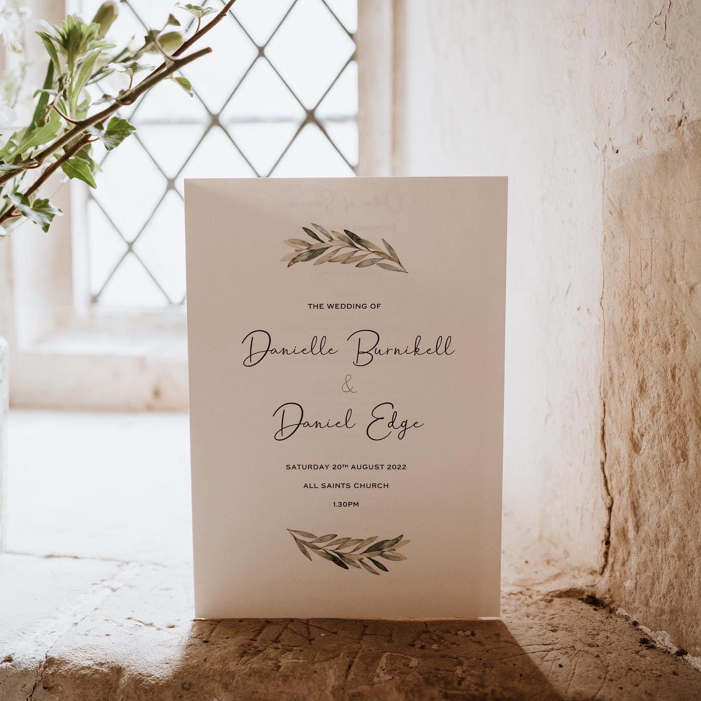Folded wedding order of service booklet with botanical illustrations, displayed upright on a rustic windowsill with soft natural light.
