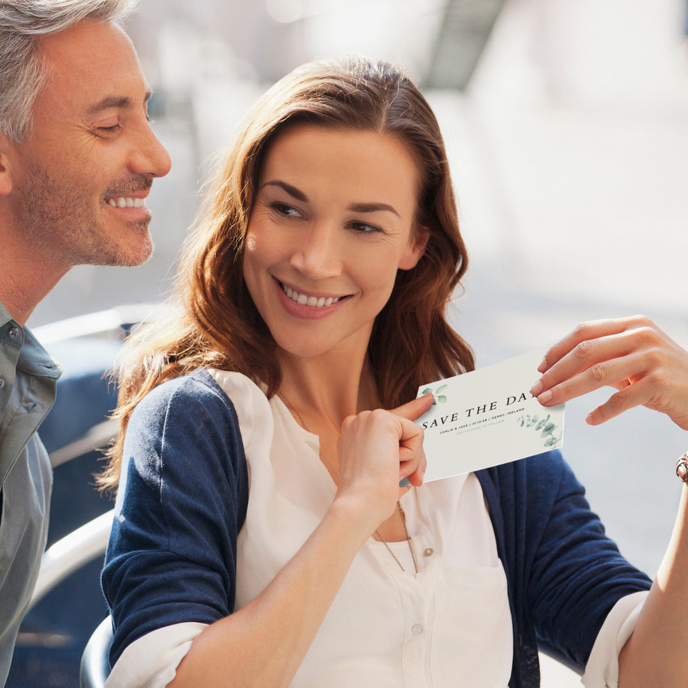 a woman in an outdoor setting, proudly holding a save the date card, showing it to a grey haired man she is sat with. They are both smiling. 
