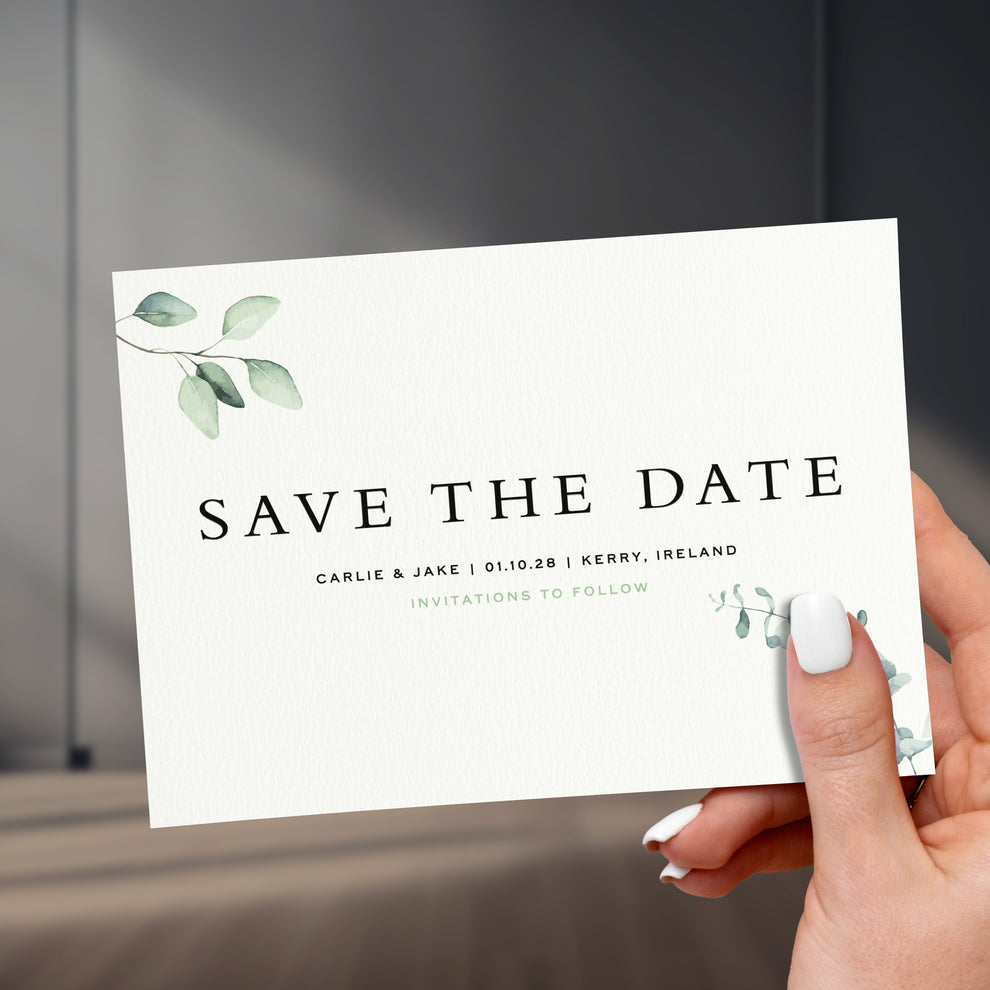 eucalyptus branch save the date card held in a hand with painted white names, held up against a stylish sleek room background