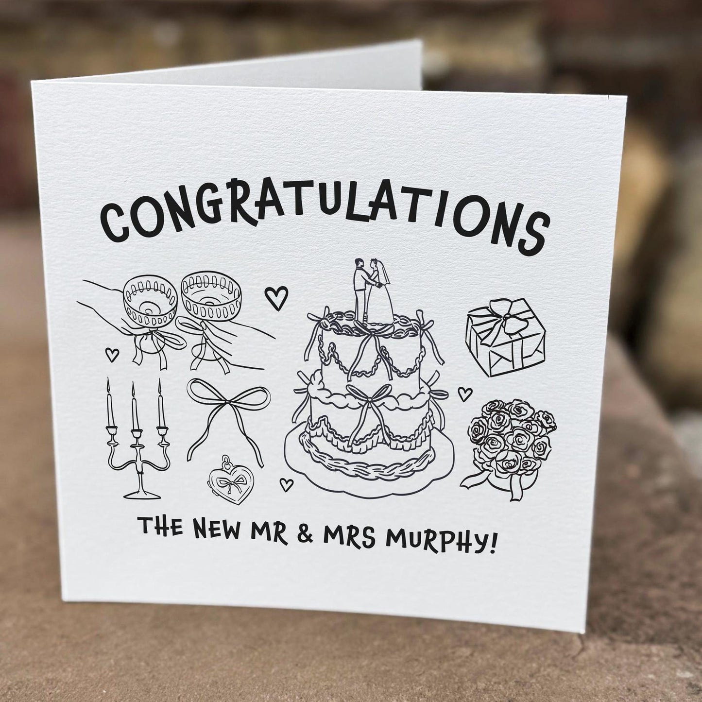 Wedding congratulations card featuring a hand-drawn cake, rings and bouquet with the text ‘Congratulations’.