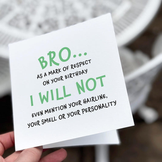 Funny bro birthday card with text ‘Bro… as a mark of respect on your birthday I will not even mention your hairline, your smell or your personality’, held in hand against white outdoor background
