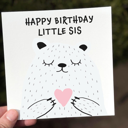 Cute illustrated birthday card showing a smiling bear holding a pink heart with the text ‘Happy Birthday Little Sis’.