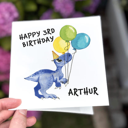 Children’s birthday card with a blue dinosaur holding balloons and wearing a party hat, text reads ‘Happy Birthday’.