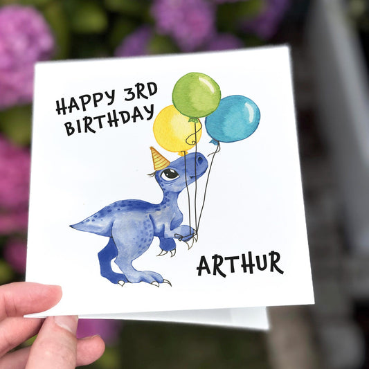 Children’s birthday card with a blue dinosaur holding balloons and wearing a party hat, text reads ‘Happy Birthday’.