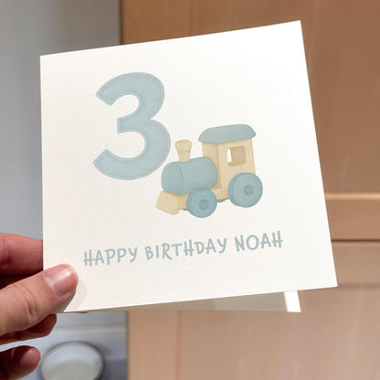 Card with a pastel blue train and large number 3, text reads ‘Happy Birthday’.