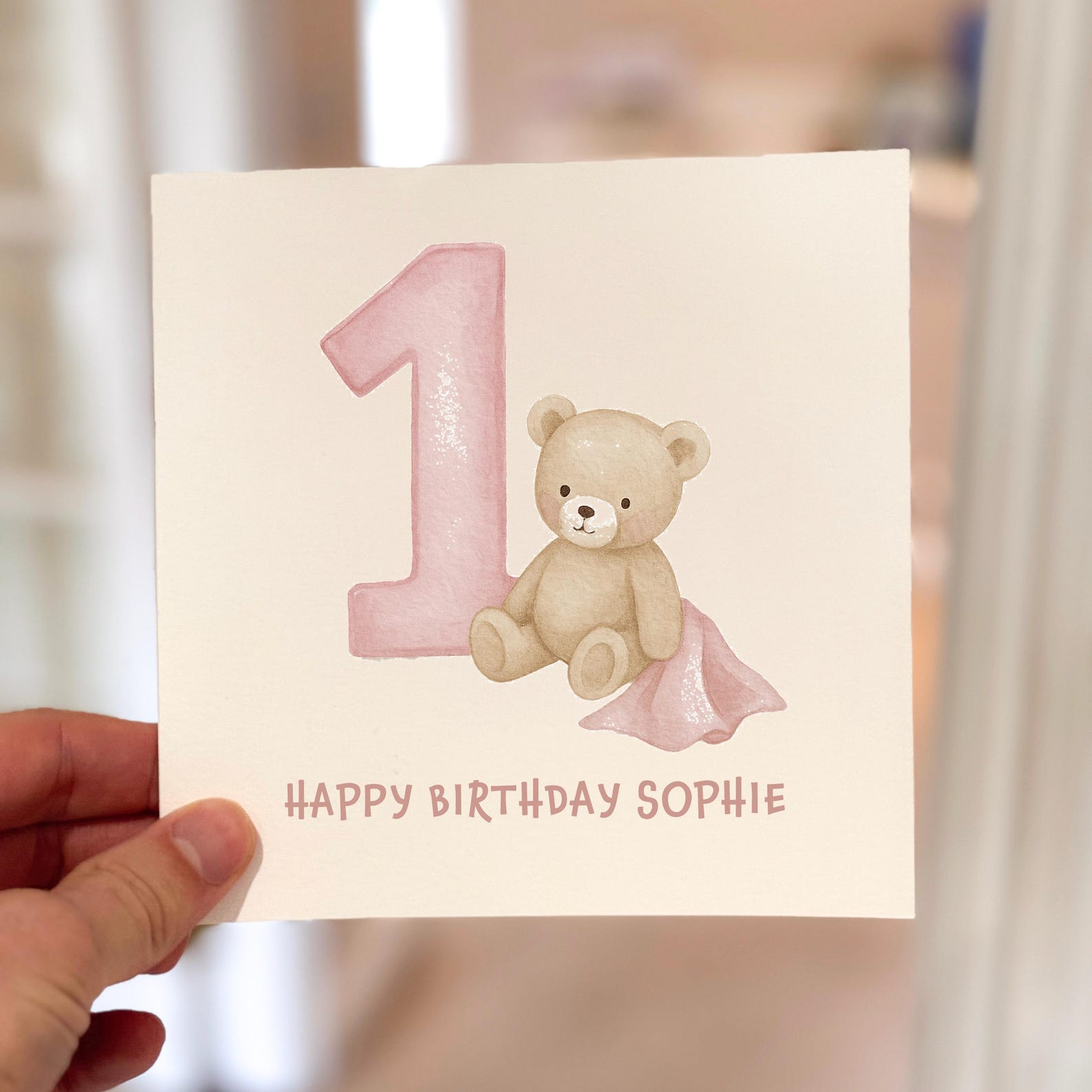 Pink 1st birthday card with teddy bear and blanket illustration, text reads ‘Happy Birthday’.