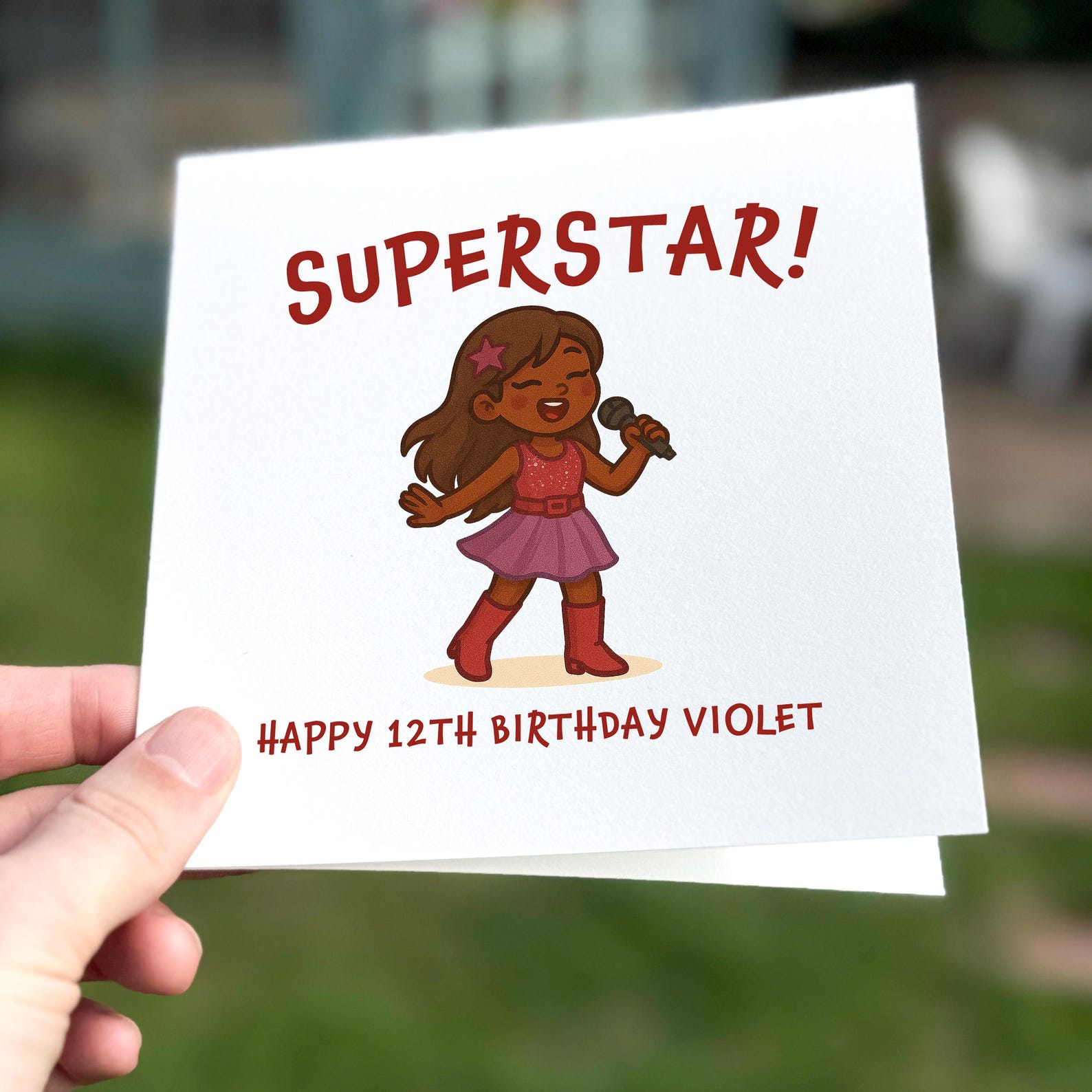 Birthday card featuring a cartoon girl singing with a microphone wearing a pink dress, text reads ‘Superstar!’.