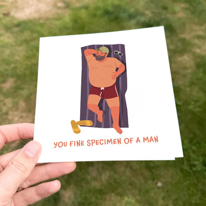 Funny birthday card showing a cartoon man sunbathing on a towel with sunglasses beside him and the text ‘You fine specimen of a man’.