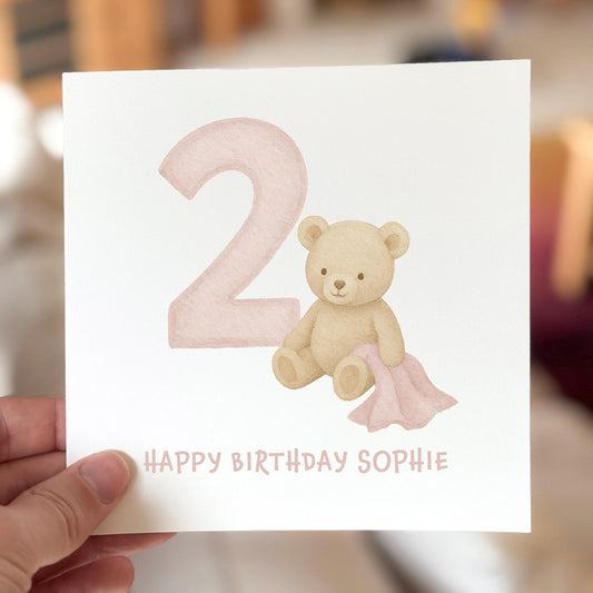 Birthday card featuring a teddy bear with a pink number two, printed on white textured card.
