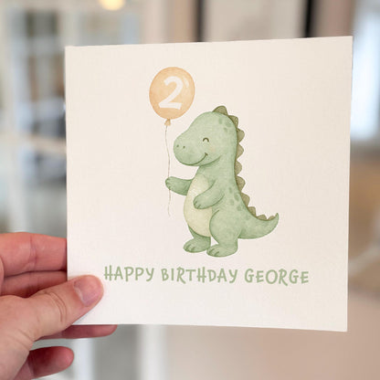 Birthday card featuring a smiling green dinosaur holding a number two balloon, printed on textured white card.