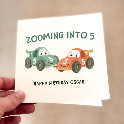 Birthday card featuring two cartoon race cars with smiling faces and the text ‘Zooming Into 5’, printed on textured card.