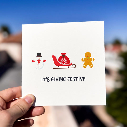 Modern festive card featuring a snowman, sleigh, and gingerbread man with the text ‘It’s Giving Festive’.