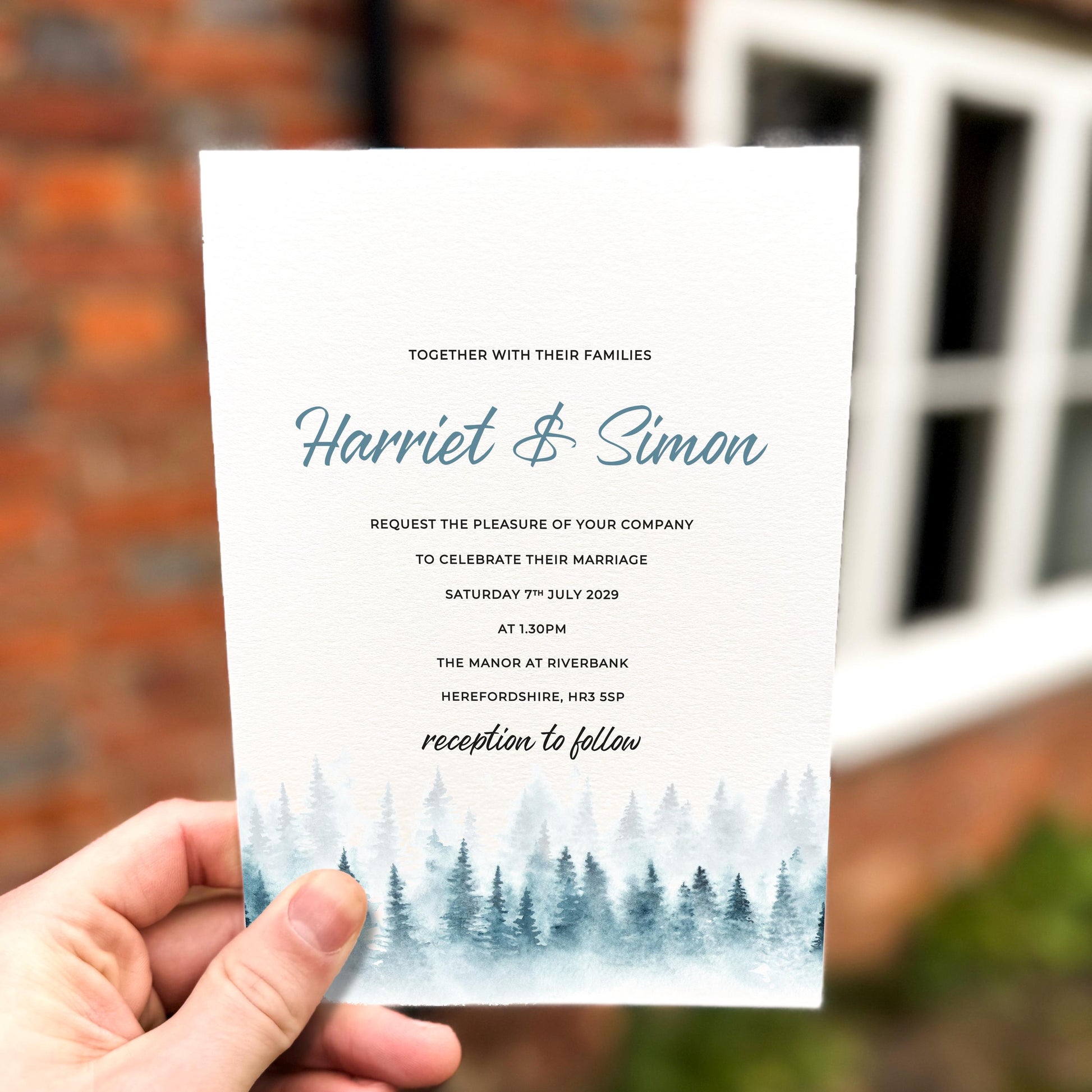 Hand holding a wedding invitation featuring soft blue watercolour pine trees along the bottom edge with elegant script text.