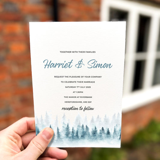 Hand holding a wedding invitation featuring soft blue watercolour pine trees along the bottom edge with elegant script text.