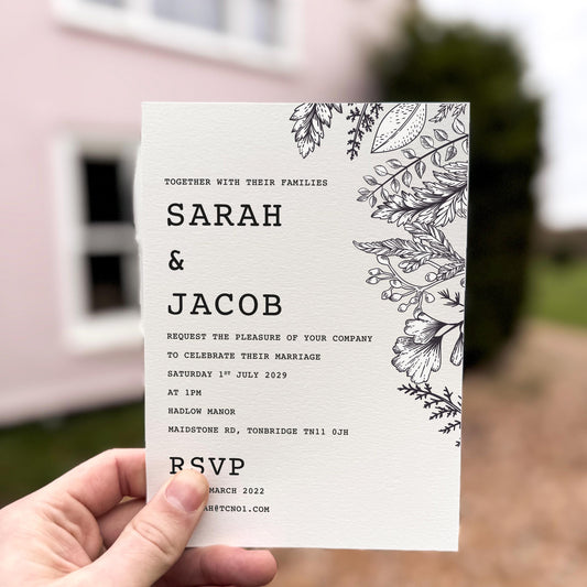 Hand holding a wedding invitation featuring detailed black botanical line art framing bold modern text.