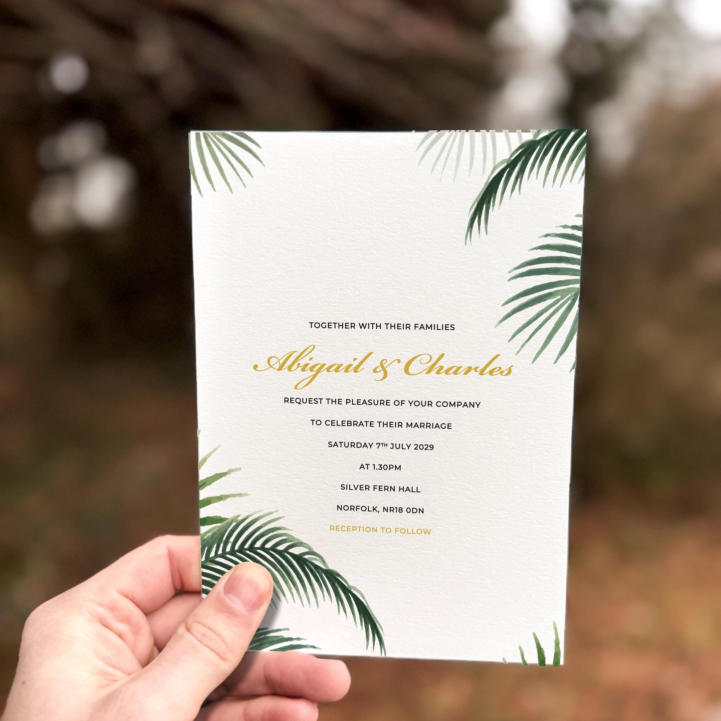 Hand holding a wedding invitation with tropical green palm leaves framing elegant gold script text.