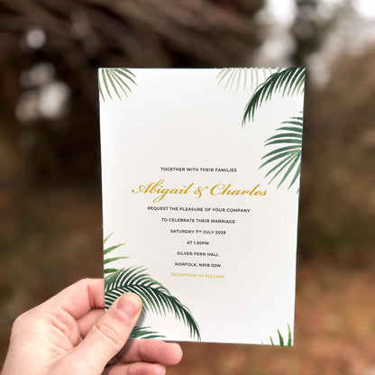 Hand holding a wedding invitation with tropical green palm leaves framing elegant gold script text.