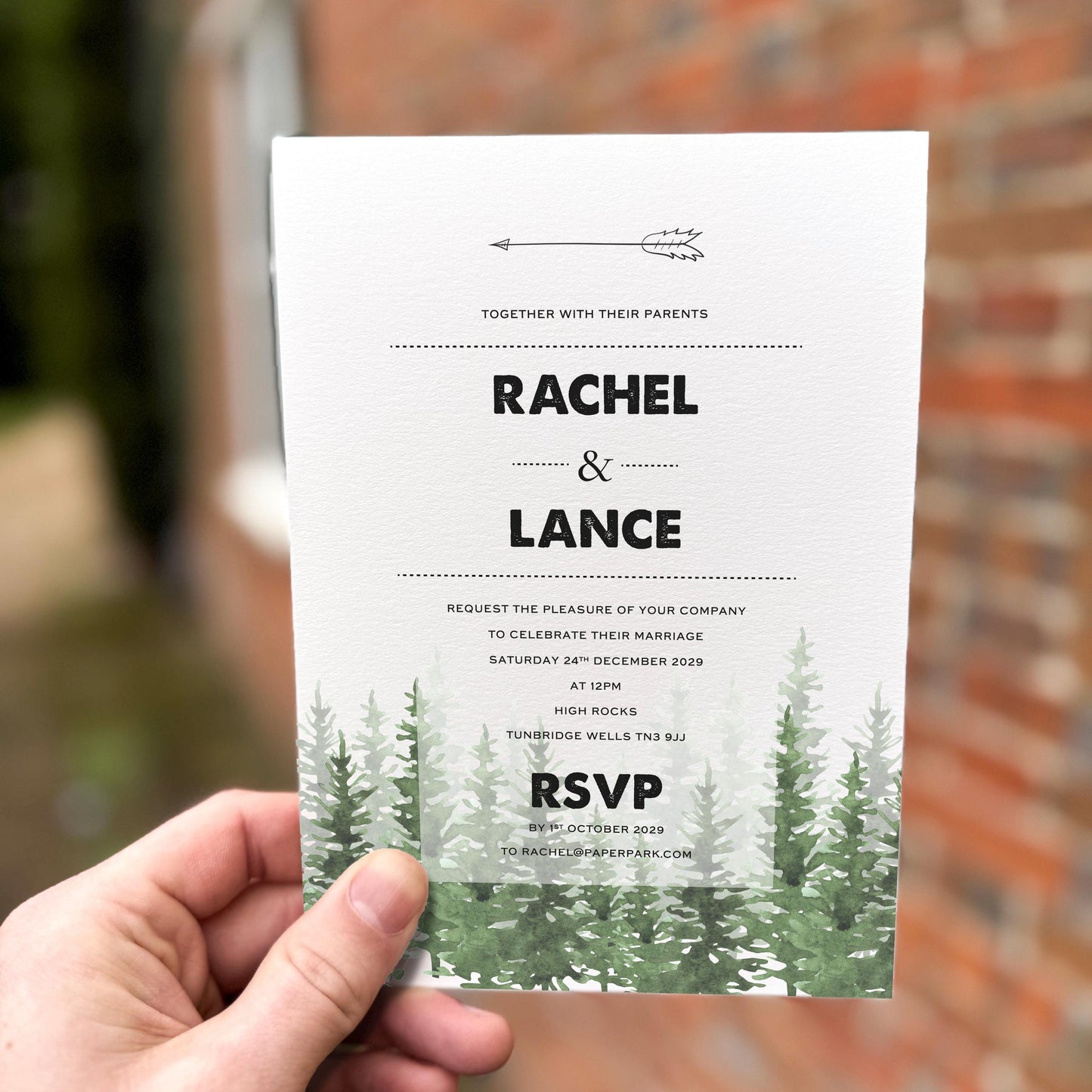 Hand holding a wedding invitation featuring a black arrow motif and green watercolour pine trees along the bottom.