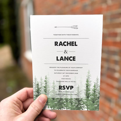 Hand holding a wedding invitation featuring a black arrow motif and green watercolour pine trees along the bottom.