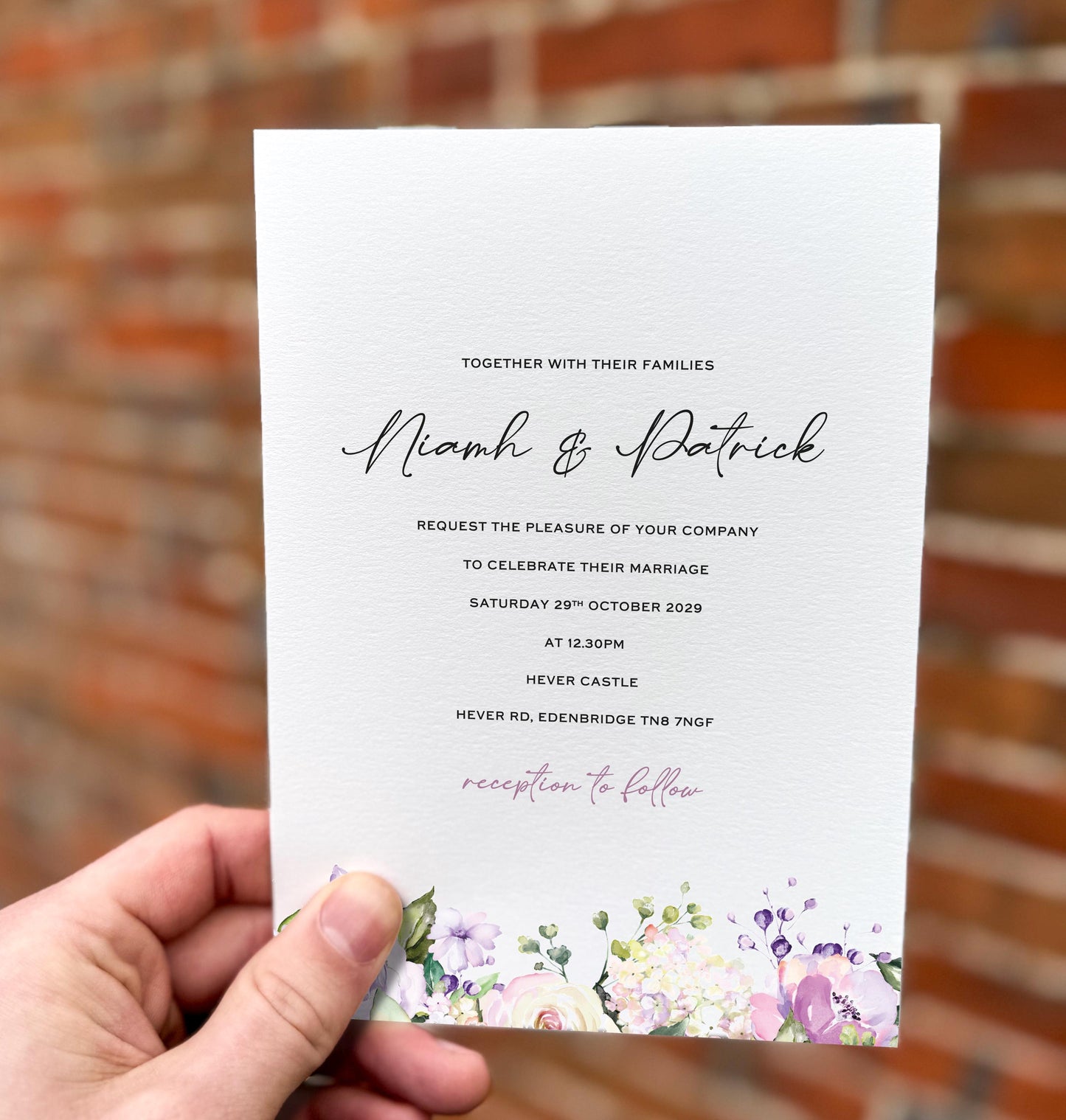 Hand holding a wedding invitation featuring pastel lilac, blush and cream florals along the bottom edge with elegant script text.