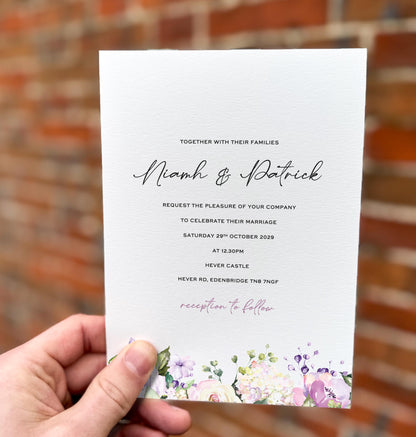 Hand holding a wedding invitation featuring pastel lilac, blush and cream florals along the bottom edge with elegant script text.