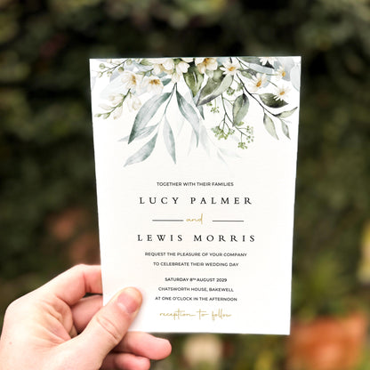 Hand holding a wedding invitation featuring soft white blossoms and flowing green foliage across the top edge.