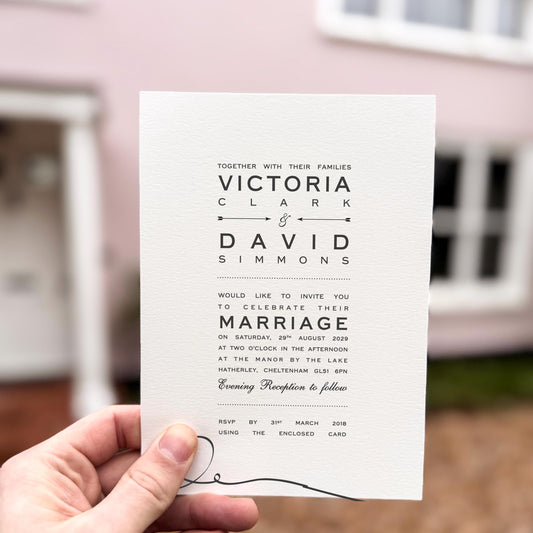 Hand holding a wedding invitation featuring bold black typography with minimalist arrows and a simple monochrome layout.