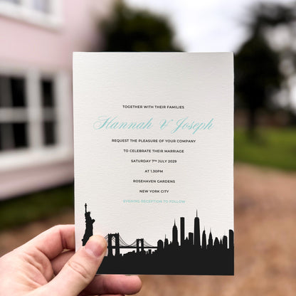 Hand holding a wedding invitation featuring a black silhouette of the New York City skyline, including the Statue of Liberty and Brooklyn Bridge.